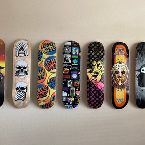 May include: Seven skateboards with various designs are displayed against a neutral wall. The boards feature graphics including skulls, a baby-like figure in a cloak, and the Santa Cruz logo. Colors include black, orange, yellow, and white.