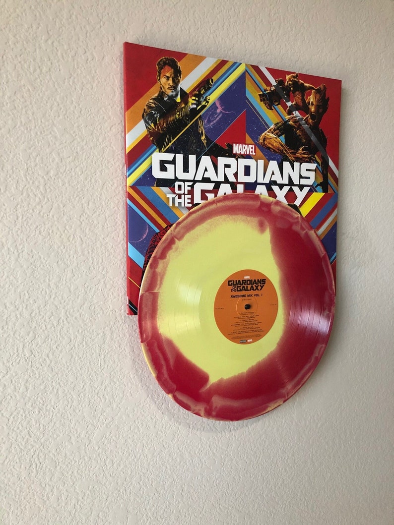 May include: A vinyl record with a yellow and red gradient design is displayed in front of a Marvel "Guardians of the Galaxy" poster. The poster features characters from the movie and the title "Guardians of the Galaxy" in white text.