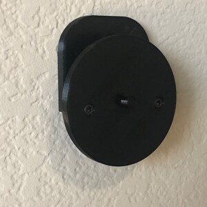 May include: A black, wall-mounted, circular object with two screws and a rectangular base. The object is made of a matte material and is mounted on a textured, off-white wall.