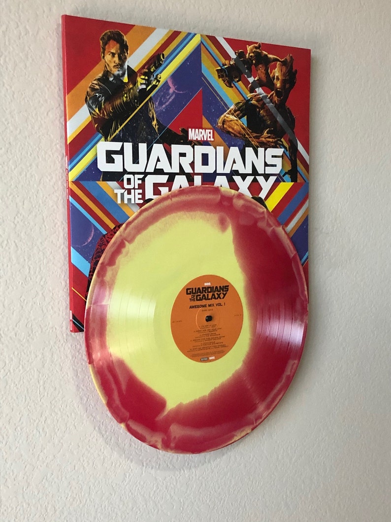 May include: A vinyl record with a yellow and red swirl design, featuring the text "Awesome Mix Vol. 1". The record is displayed in front of a Marvel "Guardians of the Galaxy" poster with character illustrations and the movie title.