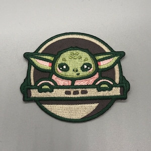 May include: Green embroidered patch of a cartoon character with large eyes and ears, wearing a pink shirt and holding a brown and green object. The character is in a circle with a brown background and a white outline.