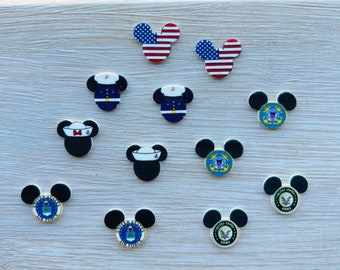 May include: A collection of Mickey Mouse-shaped pins with various designs. Some feature the American flag, while others display military emblems and uniforms. The pins are black, white, red, and blue, and are arranged on a light wooden surface.