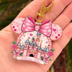 May include: A pink and white Disney Girl keychain with a gold clasp. The keychain features a castle design with a pink bow, Minnie Mouse ears, and the words "Disney Girl".