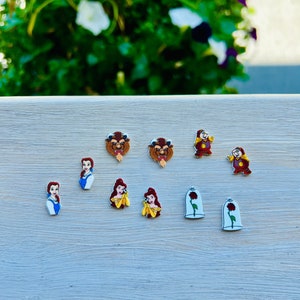 May include: A collection of colorful character charms featuring designs from the animated film, Beauty and the Beast. The charms include Belle, the Beast, Cogsworth, and the enchanted rose. The charms are arranged on a light gray surface.