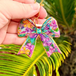 May include: A colorful bow-shaped keychain with a gold clasp. The bow features a vibrant floral pattern in shades of pink, yellow, green, and blue. The design is detailed and eye-catching, perfect for adding a touch of personality to keys or bags.