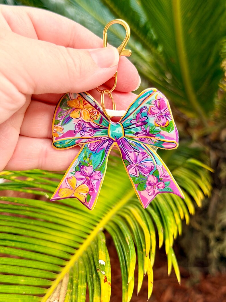 May include: A colorful bow-shaped keychain with a gold clasp. The bow features a vibrant floral pattern in shades of pink, yellow, green, and blue. The design is detailed and eye-catching, perfect for adding a touch of personality to keys or bags.