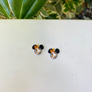 May include: Two small, round earrings featuring a Minnie Mouse design with a BB-8 droid theme. The earrings have black mouse ears, an orange bow, and a white and orange droid face. The earrings are set against a white background.