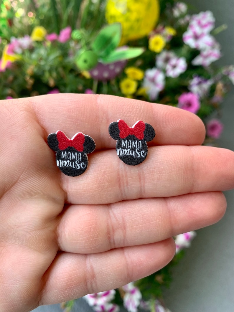 Pu&ograve; includere: Un paio di orecchini a bottone neri e rossi a forma di testa di topo con un fiocco rosso. Gli orecchini hanno la scritta "Mama Mouse" in bianco. Gli orecchini sono tenuti in una mano, con uno sfondo floreale sfocato.