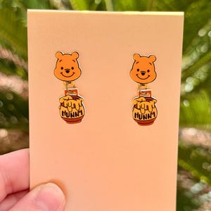 May include: A pair of Winnie the Pooh dangle earrings. Each earring features a smiling Pooh bear face above a honey pot with the word "Hunny" printed on it. The earrings are attached to a peach-colored card.