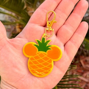 May include: A gold-colored keychain with a pineapple design. The pineapple is yellow and orange with green leaves and Mickey Mouse ears. The keychain has a gold clasp.