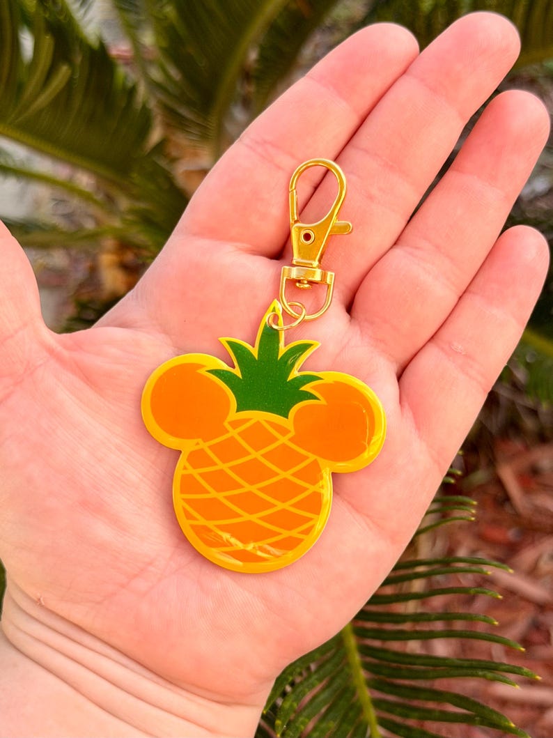 May include: A gold-colored keychain with a pineapple design. The pineapple is yellow and orange with green leaves and Mickey Mouse ears. The keychain has a gold clasp.