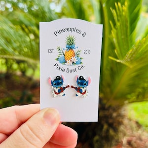 May include: A pair of Stitch-themed earrings, each depicting the character playing a red guitar. The earrings are on a white card with the text "Pineapples & Pixie Dust Co." and a pineapple graphic. The background is blurred green foliage.