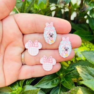 May include: Four decorative resin embellishments. Two are shaped like Minnie Mouse heads with pink gingham patterns and bows. The other two are oval-shaped with Minnie Mouse illustrations, surrounded by a light blue and pink design.
