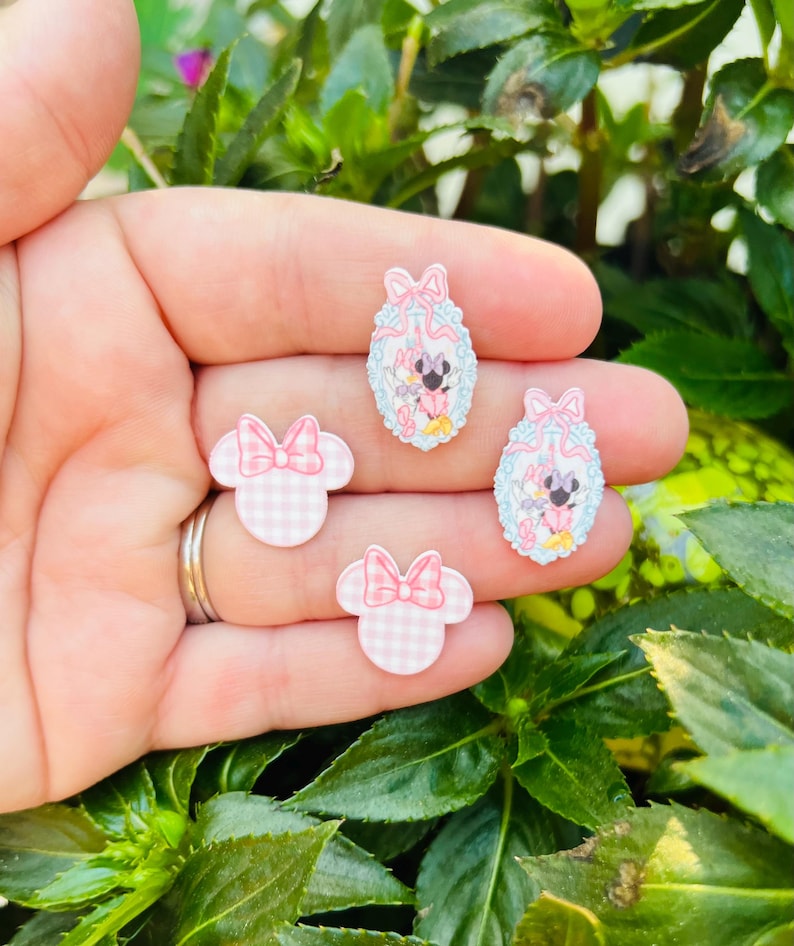 May include: Four decorative resin embellishments. Two are shaped like Minnie Mouse heads with pink gingham patterns and bows. The other two are oval-shaped with Minnie Mouse illustrations, surrounded by a light blue and pink design.