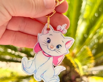 May include: A white cat keychain with a pink bow and collar, and blue eyes. The cat is sitting with its paws forward. The keychain has a gold-colored clasp.