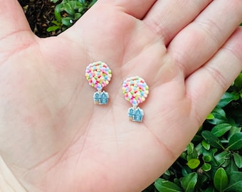 May include: A pair of whimsical stud earrings. Each earring features a colorful cluster of tiny beads resembling balloons, attached to a miniature house charm. The earrings are held in a hand against a backdrop of green foliage.