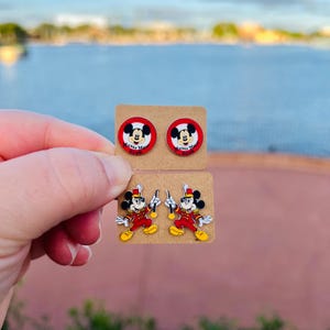 Op de afbeelding: Twee paar Mickey Mouse-oorbellen. E&eacute;n paar heeft Mickey's gezicht in een rode cirkel met de tekst "Mickey Mouse Club". Het andere paar toont Mickey in een rood-geel uniform, met een baton. Beide paren zitten op bruine oorbellenkaarten.
