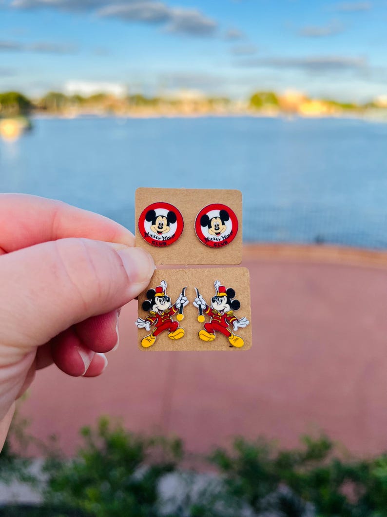 Op de afbeelding: Twee paar Mickey Mouse-oorbellen. E&eacute;n paar heeft Mickey's gezicht in een rode cirkel met de tekst "Mickey Mouse Club". Het andere paar toont Mickey in een rood-geel uniform, met een baton. Beide paren zitten op bruine oorbellenkaarten.