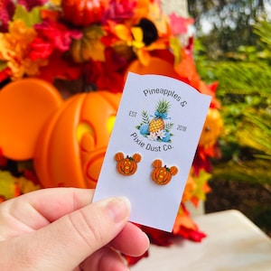 May include: A pair of pumpkin Mickey Mouse earrings on a white card with the text "Pineapples & Pixie Dust Co. Est 2018".
