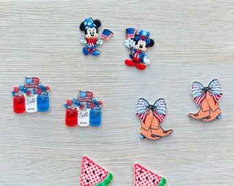 Può includere: Una collezione di ciondoli e abbellimenti a tema patriottico. Include personaggi di Topolino, barattoli Mason con bandiere, stivali da cowboy con fiocchi e fette di anguria. I ciondoli sono rossi, bianchi, blu e verdi.