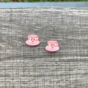 May include: Two pink teacup and saucer decorations. The teacups have a heart design in the center and are trimmed with gold. The saucers are also pink and have a heart design. The teacups and saucers are small and decorative.