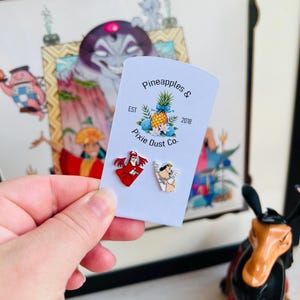 May include: A person holding a white card with two character pins and the text "Pineapples & Pixie Dust Co.". The pins are of a red character and a tan character with wings. The background features a framed illustration.