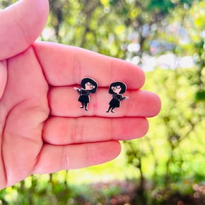 May include: A pair of black and silver stud earrings featuring a cartoon character design. The earrings depict a woman with black hair, glasses, and a black outfit. The earrings are small and detailed, perfect for fans of the animated film.