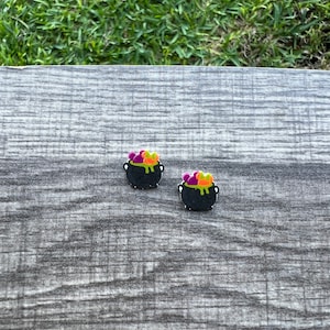 May include: Two black cauldron-shaped earrings with colorful bubbling potion. The potion is a mix of purple, orange, and green. The earrings are set against a gray wooden surface with green grass in the background.