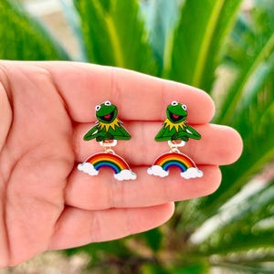 May include: A pair of earrings featuring a green frog character with a yellow collar and a rainbow. The earrings are attached to a gold-colored hook. The background is a blurred image of green leaves.