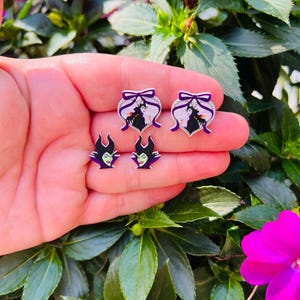 Puede incluir: Un conjunto de cuatro pendientes con el personaje Mal&eacute;fica de la pel&iacute;cula de Disney La Bella Durmiente. Los pendientes son de plata con detalles en morado y negro. Dos pendientes muestran la cara de Mal&eacute;fica, y dos pendientes muestran a Mal&eacute;fica en su forma de drag&oacute;n.