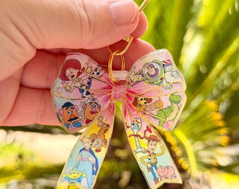 May include: A pink and gold bow-shaped keychain featuring characters from the animated film Toy Story. The keychain has a gold clasp and a pink ribbon design with images of Woody, Buzz Lightyear, and other characters.