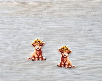 May include: Two brown Highland cow stickers, each wearing a sunflower crown. The stickers are sitting on a light wood-grain surface, with green grass visible in the background. The cows are in a sitting position.