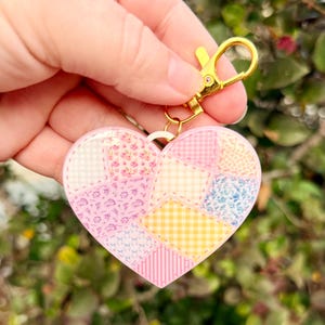 May include: Heart-shaped keychain with a patchwork design. The keychain features a gold-colored clasp and ring. The heart is made of pink acrylic and has a patchwork design with various floral and gingham patterns in pastel colors.