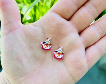 Op de afbeelding: Twee kleine, kleurrijke oorbellen met een theekopjesontwerp met een konijn erin. De oorbellen zijn voornamelijk rood, wit en geel, met een hartvormig detail. De oorbellen worden in een hand gehouden tegen een vage groene achtergrond.