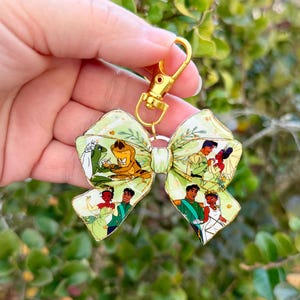 May include: A green bow-shaped keychain with gold hardware. The bow features illustrations of characters from a fairy tale, including a frog, a princess, and a prince. The background is a light green with floral accents.