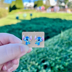 May include: A pair of blue and white Stitch stud earrings on a small, rectangular, tan card. The earrings feature the character Stitch from the Disney movie Lilo & Stitch. The background is a blurred outdoor scene with green foliage and a blue sky.