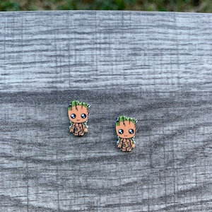 May include: Two small, colorful Groot character earrings. Each earring features a detailed design of the character's face and body, with brown and green accents. The earrings are set against a gray, textured surface.