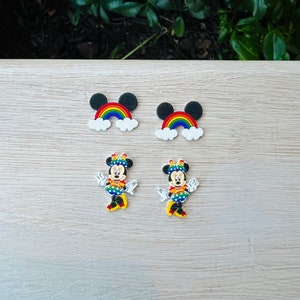 May include: Four colorful charms. Two charms feature a rainbow with black Mickey Mouse ears and white clouds. The other two charms are Minnie Mouse, wearing a rainbow polka dot dress, a matching bow, and yellow shoes.
