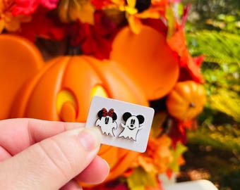 May include: A pair of Halloween-themed earrings featuring ghost versions of a mouse character with a red bow and a mouse character. The earrings are held in front of a pumpkin and fall foliage.