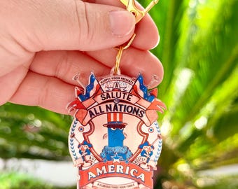 May include: A novelty keychain featuring a cartoon character in a top hat, with the text "A Salute to All Nations But Mostly America". The design incorporates red, white, and blue patriotic elements, with a gold-colored clasp.