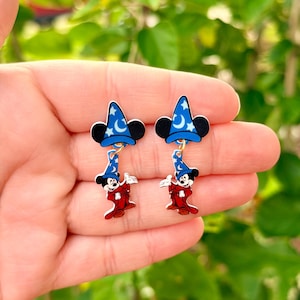 May include: A pair of earrings featuring a cartoon character in a red outfit and a blue wizard hat with white stars and a crescent moon. The earrings have black Mickey Mouse ears and are hanging from a gold-colored hook.