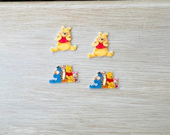 May include: Four colorful character embellishments featuring Winnie the Pooh and friends. Two show Pooh Bear in a seated pose, wearing a red shirt. The other two show Pooh, Eeyore, Piglet, and Tigger together. These are likely for crafting.