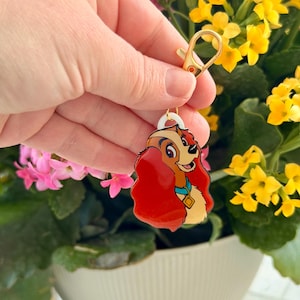May include: A gold-colored keychain with a cartoon image of a dog with red hair, a blue collar, and a gold tag. The keychain is attached to a gold clasp. The background features yellow and pink flowers in a white pot.