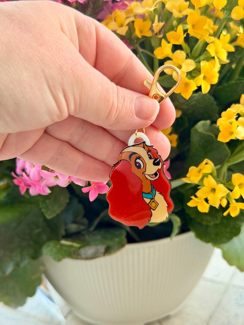 May include: A gold-colored keychain with a cartoon image of a dog with red hair, a blue collar, and a gold tag. The keychain is attached to a gold clasp. The background features yellow and pink flowers in a white pot.