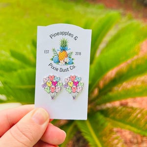 May include: A pair of heart-shaped balloon earrings in pastel colors, held against a white card with the text "Pineapples & Pixie Dust Co.". The card also features a pineapple illustration. The earrings are a fun and whimsical accessory.