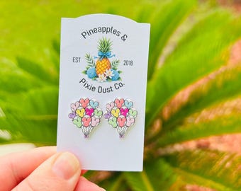 May include: A pair of heart-shaped balloon earrings in pastel colors, held against a white card with the text "Pineapples & Pixie Dust Co.". The card also features a pineapple illustration. The earrings are a fun and whimsical accessory.