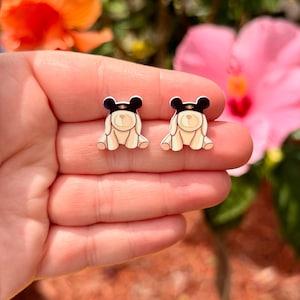 May include: A pair of cream-colored bear-shaped stud earrings with black hats and gold accents. The earrings are small and detailed, with a cartoonish design. The background features colorful flowers.
