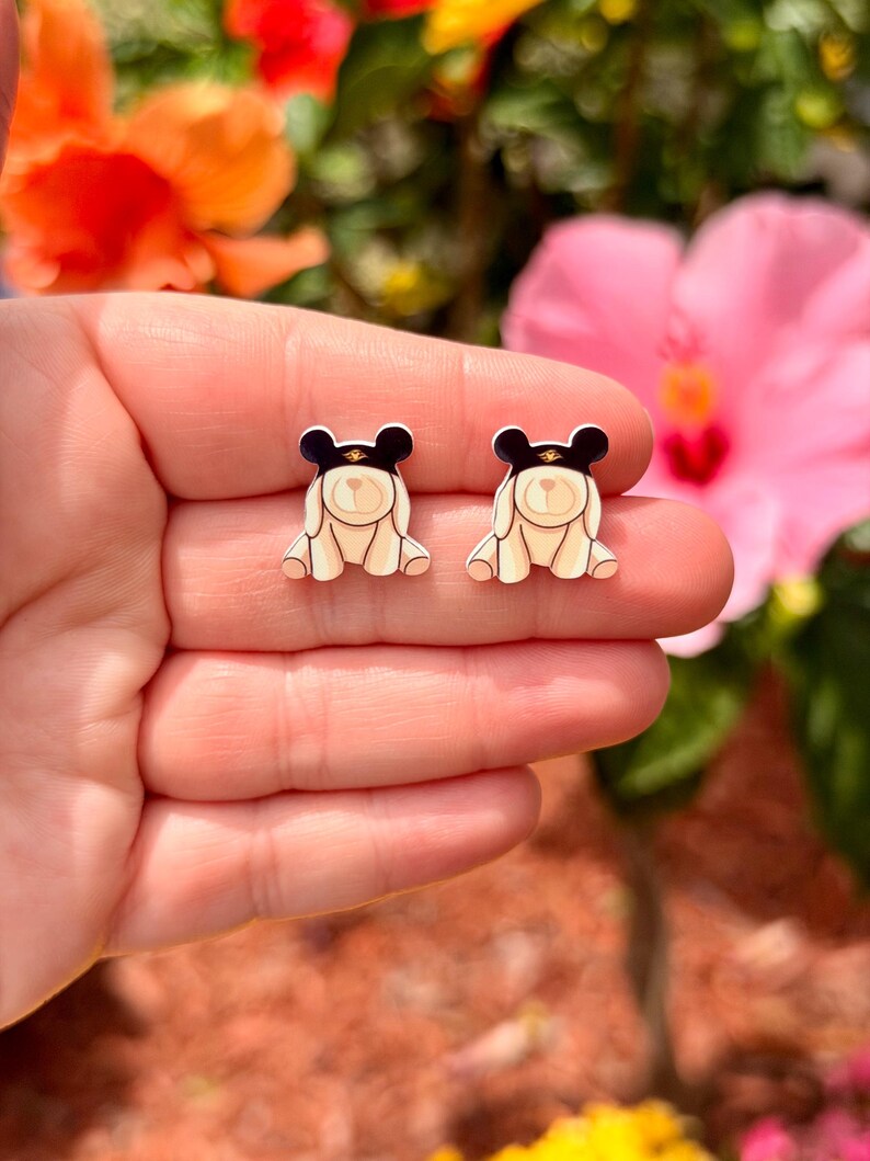 May include: A pair of cream-colored bear-shaped stud earrings with black hats and gold accents. The earrings are small and detailed, with a cartoonish design. The background features colorful flowers.