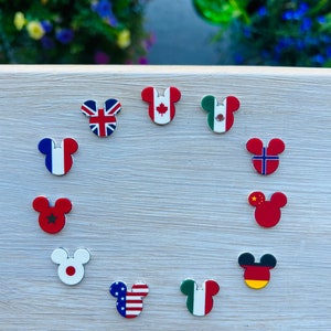 May include: A selection of Mickey Mouse-shaped magnets, each displaying a national flag. Flags include the UK, Canada, Mexico, France, Norway, China, Germany, Japan, the USA, and Italy. The magnets are arranged on a light-coloured wooden surface.