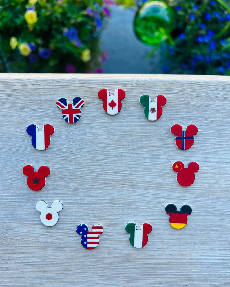 May include: A selection of Mickey Mouse-shaped magnets, each displaying a national flag. Flags include the UK, Canada, Mexico, France, Norway, China, Germany, Japan, the USA, and Italy. The magnets are arranged on a light-coloured wooden surface.
